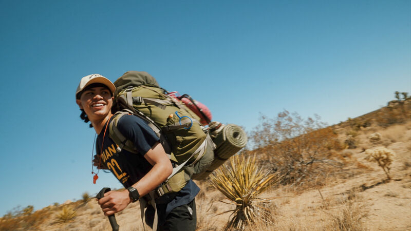 A young man is hiking in a desert landscape. He is wearing a baseball cap, a t-shirt, and a large backpack. He is using a walking stick to help him navigate the terrain. The sky is blue and clear, and the sun is shining brightly. The landscape is arid and sparsely vegetated, with cacti and scrub brush dotting the hillsides. The man appears to be enjoying his hike, as he is smiling and looking around at the scenery.